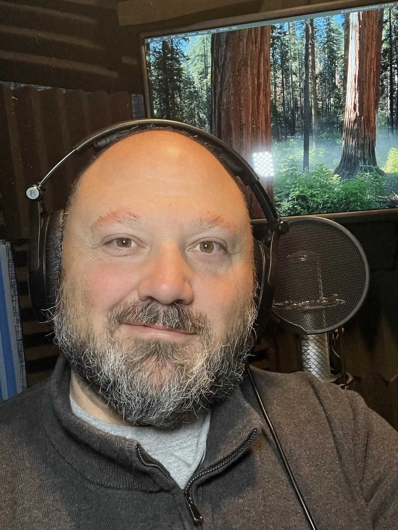 Anthony Pica smiling in his vocal booth, wearing headphones, with his Manley tube microphone and script screen behind him
