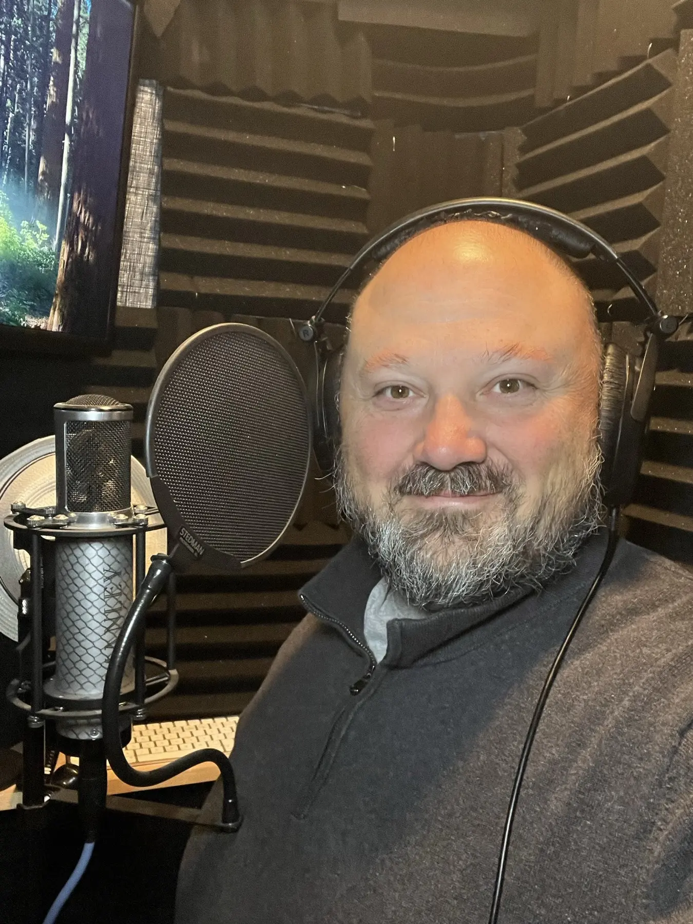 Anthony Pica smiling beside his Manley tube microphone inside his custom-built vocal booth, acoustic foam on the walls